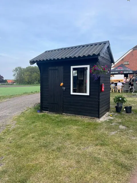 Zwart houten sanitairhuisje met bloembakken aan de rand van het veld